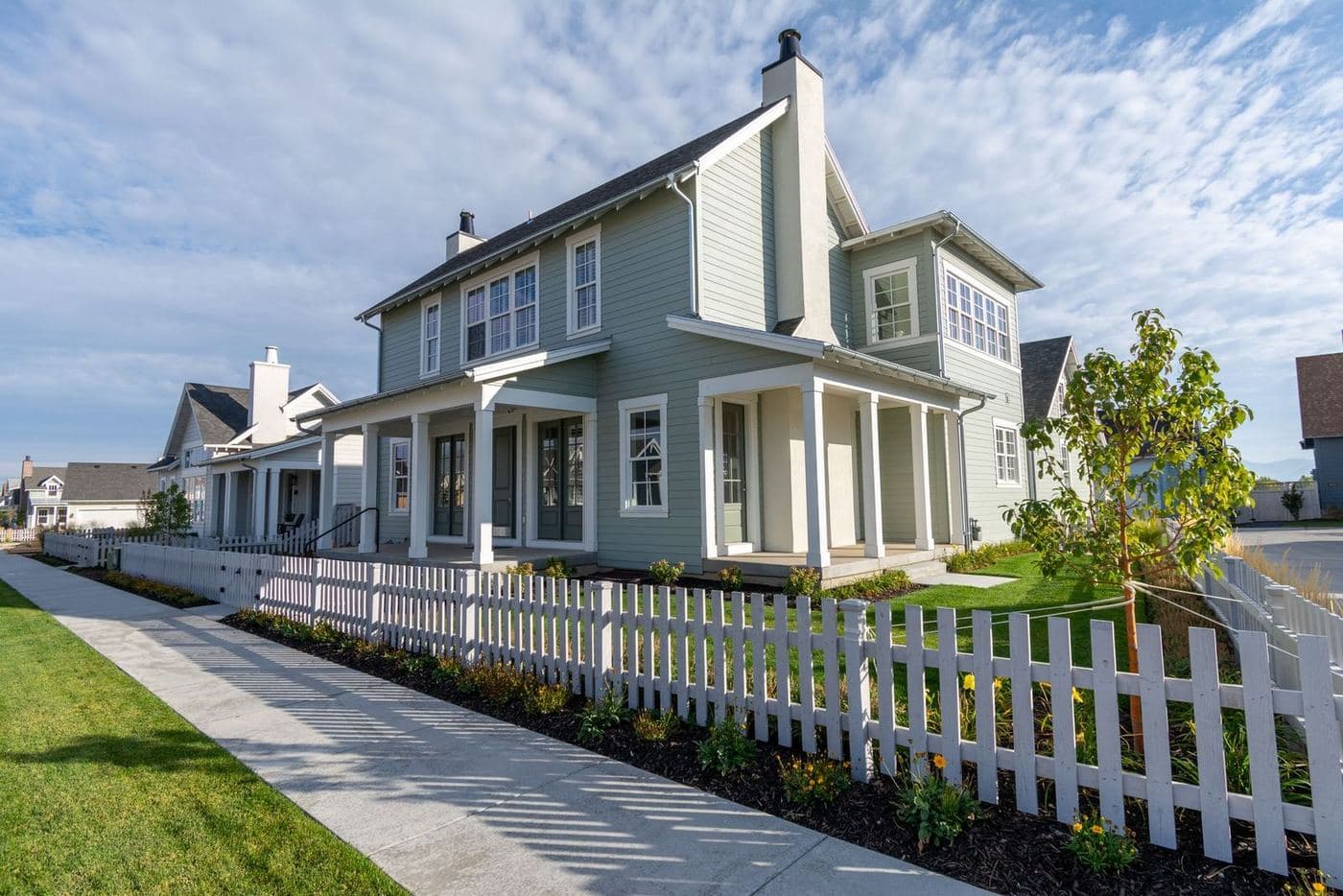 Gray two-story home with a wraparound porch behind a white picket fence