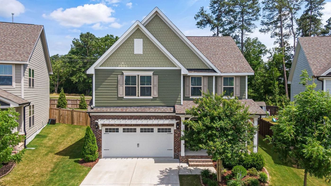 Sage-green two-story craftsman-style home with a front-load garage and young trees
